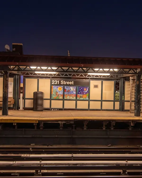 An elevated subway platform at dusk, lit under its canopy and empty, seen from across the tracks.